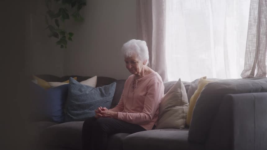 An elderly woman with gray hair sits on a couch around midday holding her hands together with a smile on her face