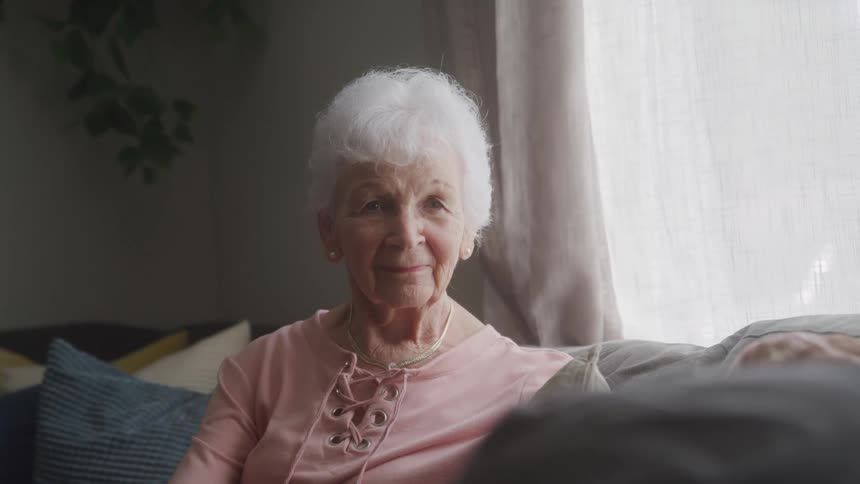 An elderly woman sits on the couch slowly turning her head in a deliberate motion