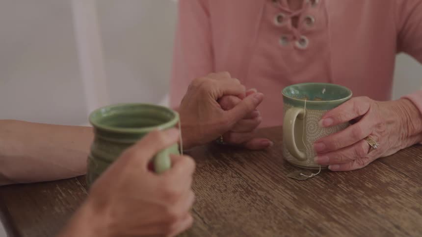Two people sit together holding warm mugs and hands in a comforting intimate moment