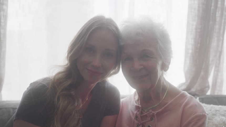 Grandmother and granddaughter sit close smiling in soft window light