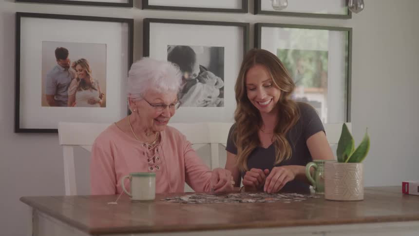 An elderly woman and a young woman sit smiling at a table while working on a puzzle together