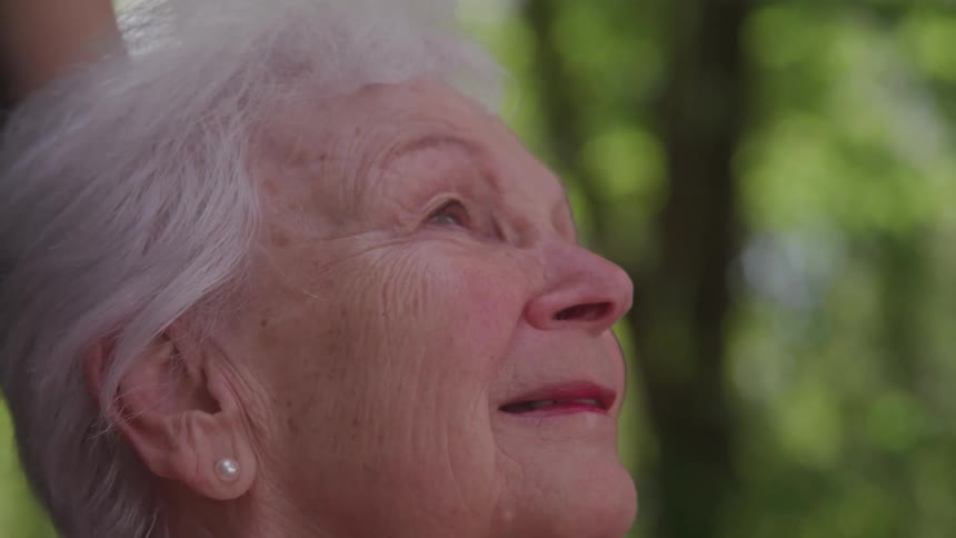 An elderly woman smiles softly while gazing upward in a peaceful outdoor setting