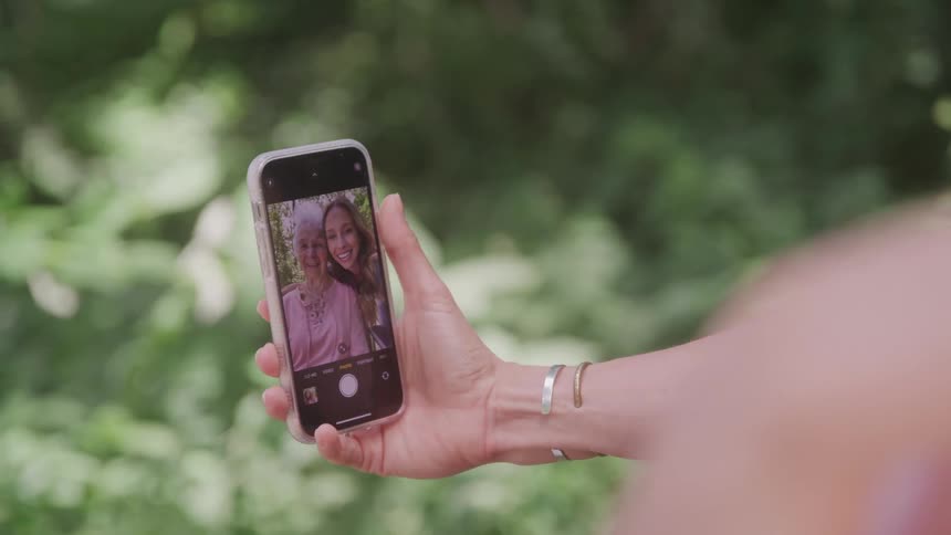 A person takes a cheerful selfie with a friend using a smartphone in a green outdoor setting