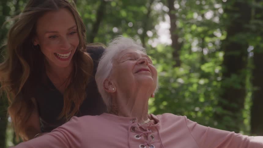 A young woman pushes an older woman in a wheelchair as they enjoy a fun time together in the woods around midday