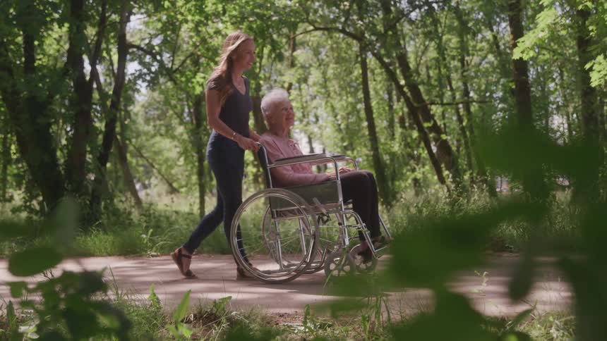A woman pushes an older woman in a wheelchair along a dirt path surrounded by tall trees and lush foliage
