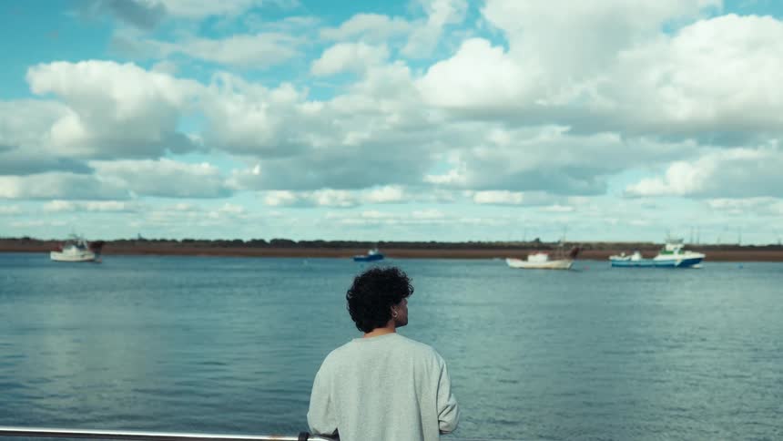 A man quietly gazes across a calm body of water with boats under a partly cloudy sky
