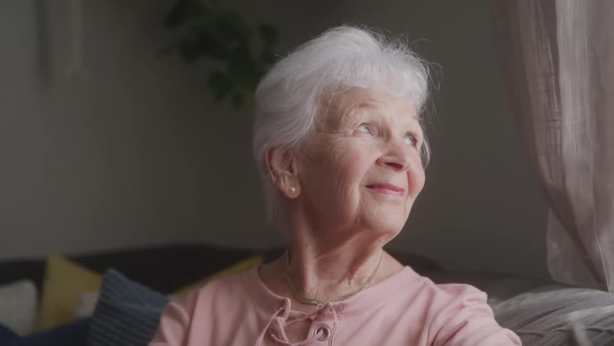 Close up portrait of an elderly woman looking out the window and smiling while sitting on a couch in a living room during daytime