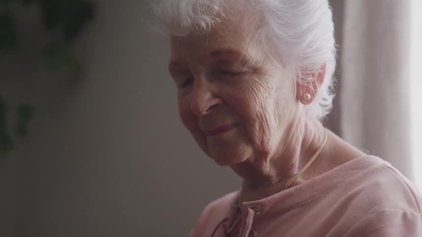 An elderly woman stands beside a window around midday with a smile on her face