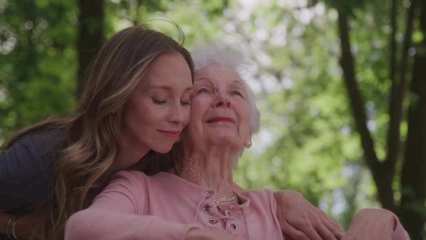 Young woman hugs elderly woman from behind in a peaceful outdoor moment