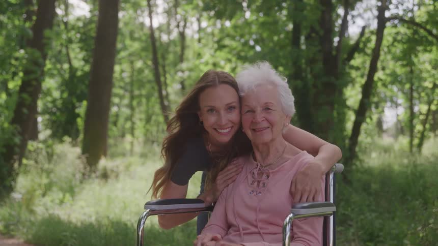 A young woman hugs an elderly woman in a wheelchair as they smile together in a sunlit forest