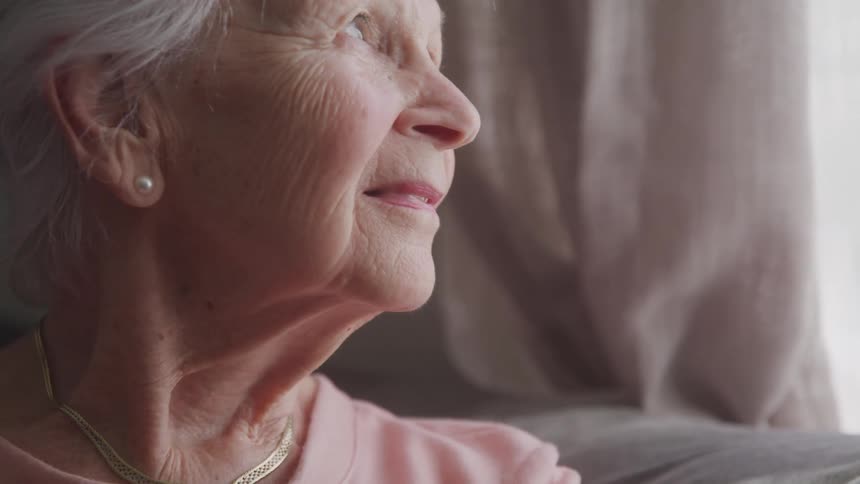 An elderly person with white hair wears a pink shirt pearl earring and gold necklace while seated indoors beside a curtained window
