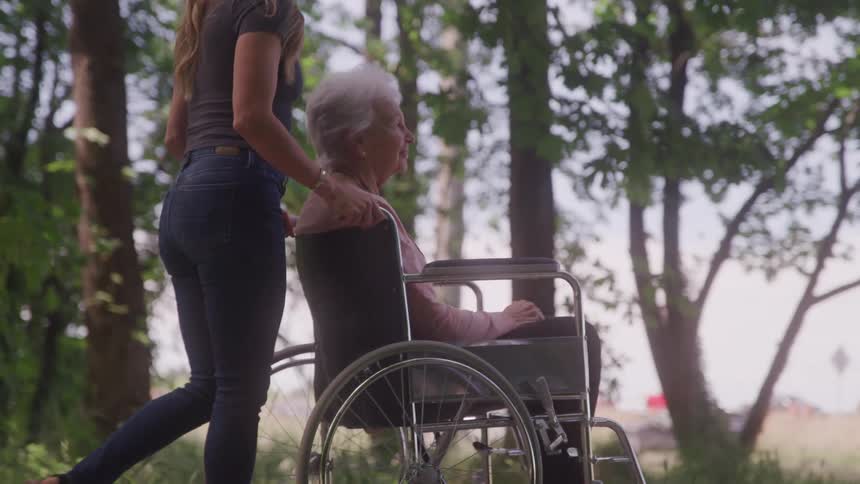 A woman is pushing an elderly woman in a wheelchair through a forested area