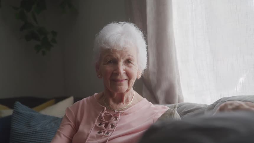 An elderly woman sits on a couch smiling warmly at the camera in a softly lit room