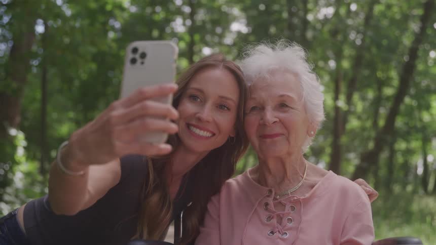 A young woman and an elderly woman are smiling while taking a selfie together outdoors