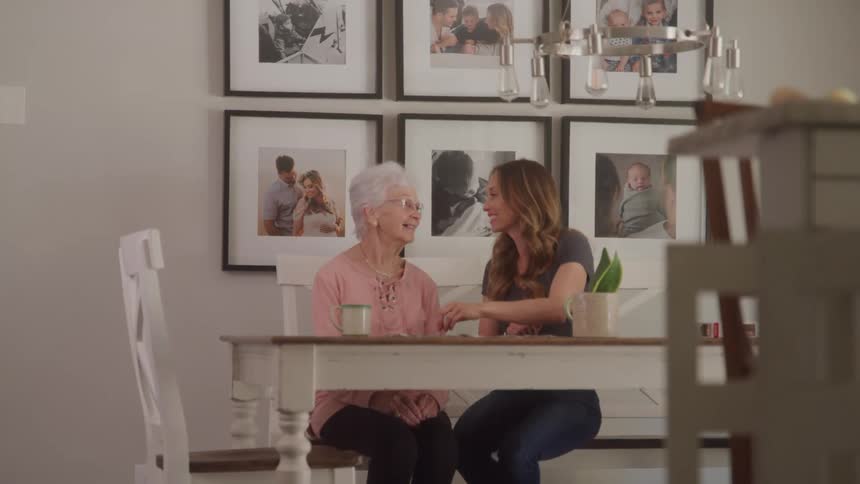 An elderly woman and a younger woman sit together at a dining table smiling and chatting in a room decorated with family photos
