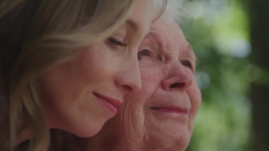 An elderly woman sits in a public park with her daughter standing behind her both smiling