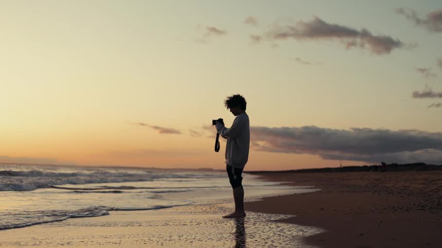 Silhouette of a man in a white shirt and a professional camera on the sand of a beach at dawn