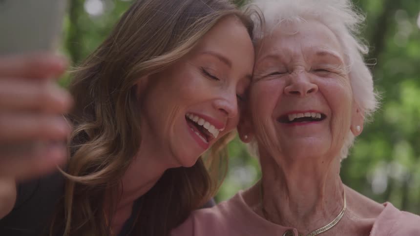 A young woman and an elderly woman smile closely together while taking a selfie outdoors
