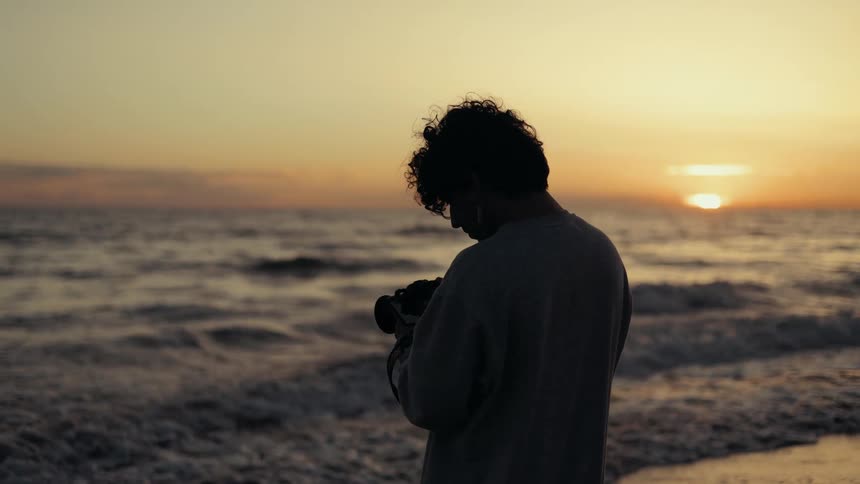 A person stands on the beach at sunset looking down at the camera