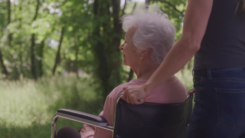 Close up of a young woman pushing an elderly woman on a wheelchair along a path in the middle of a woodland area during daytime