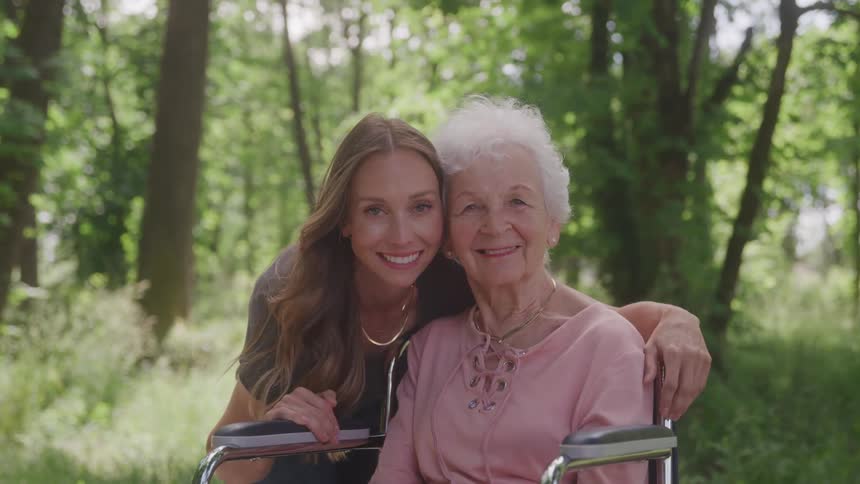 A young woman lovingly hugs an elderly woman in a wheelchair outdoors