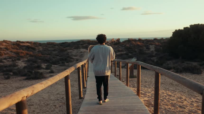 A person stands on a wooden walkway leading through a sandy and rocky landscape toward the ocean at sunset