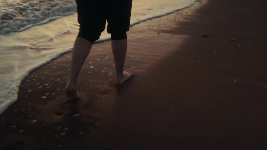 Man walking barefoot across the beach at sunset carries a camera