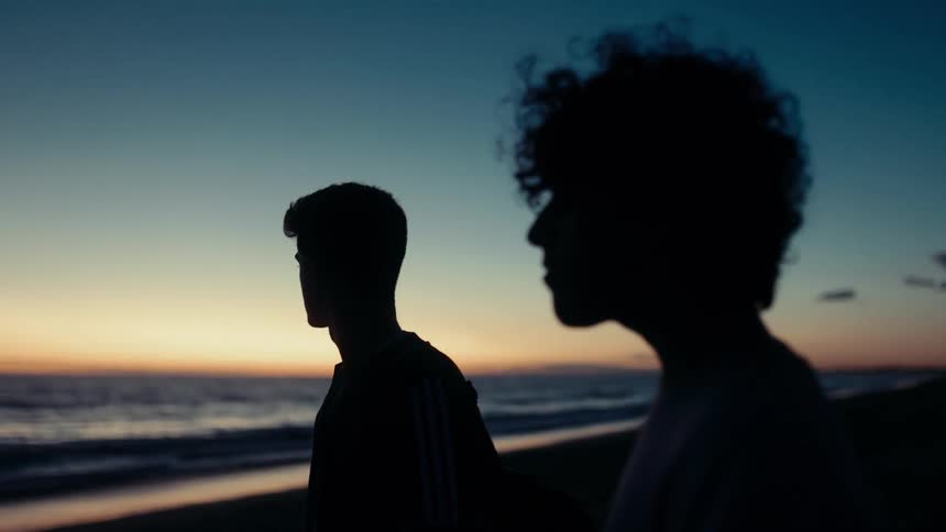 Two silhouetted teenagers stand on a beach at sunset framed by a gradient sky and calm ocean waves