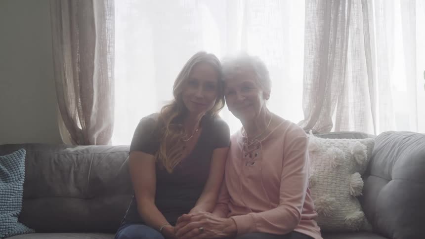 Two women sit close on a couch smiling and holding hands