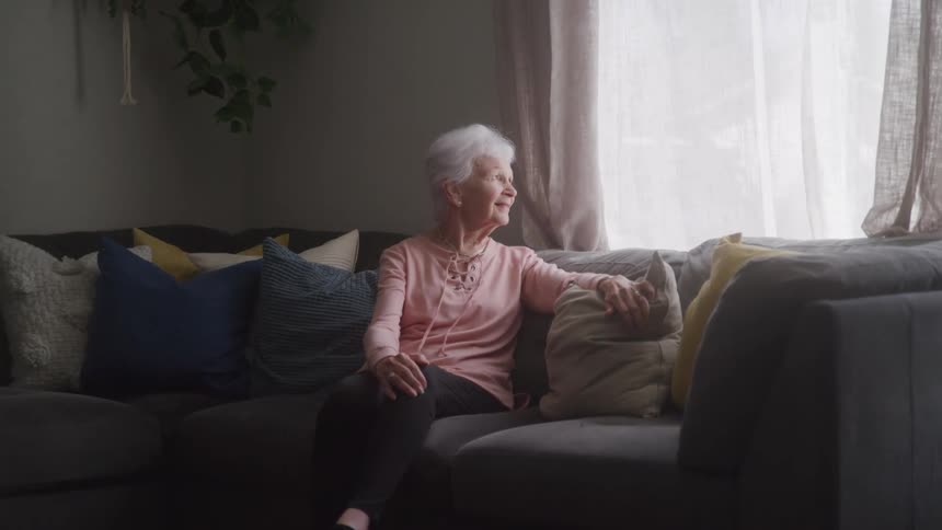 Elderly woman sits on couch gazing out window thoughtfully