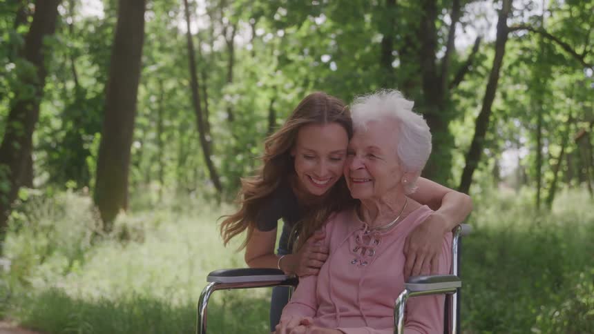 An elderly woman sits in a wheelchair as her daughter stands behind her hugging her in a public park around midday