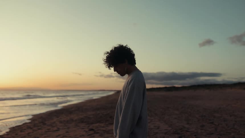 A teenager stands alone on a sandy beach at sunset looking out towards the ocean with his profile silhouetted against the horizon