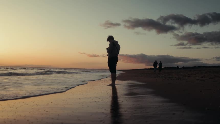 A man stands still on the beach as ocean waves hit the sand with the sun setting in the background