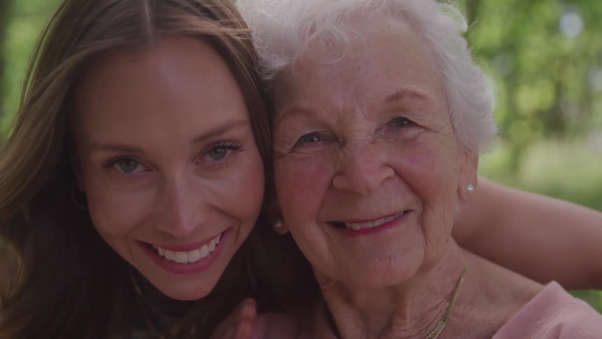 A young woman hugs an elderly woman from behind in a public park around midday