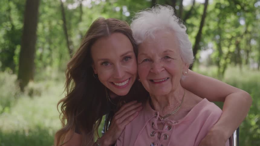 A young woman with long hair embraces an older woman with white hair both smiling directly at the viewer with a green leafy background