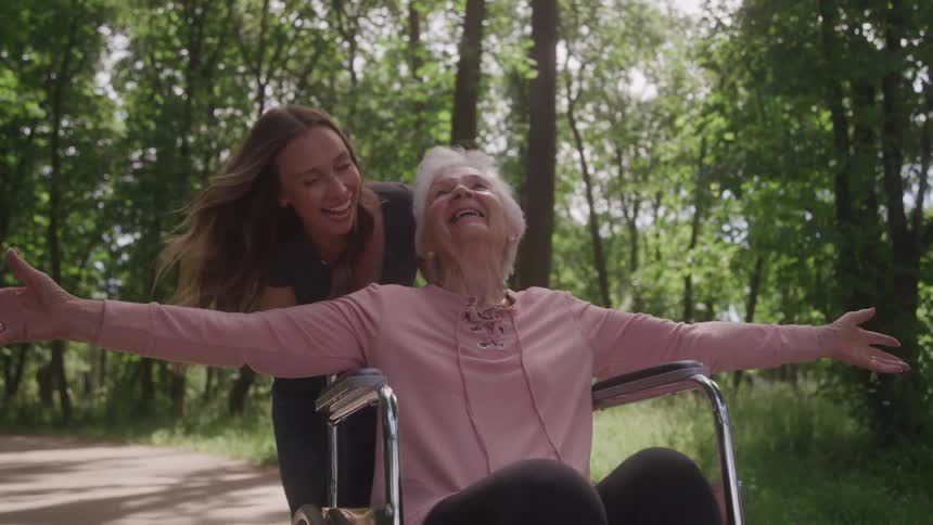 Elderly woman in wheelchair is joyfully pushed by younger woman on sunny forest path