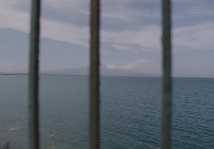 The view through vertical metal bars shows a calm blue ocean stretching towards a distant landmass under a sky with scattered clouds