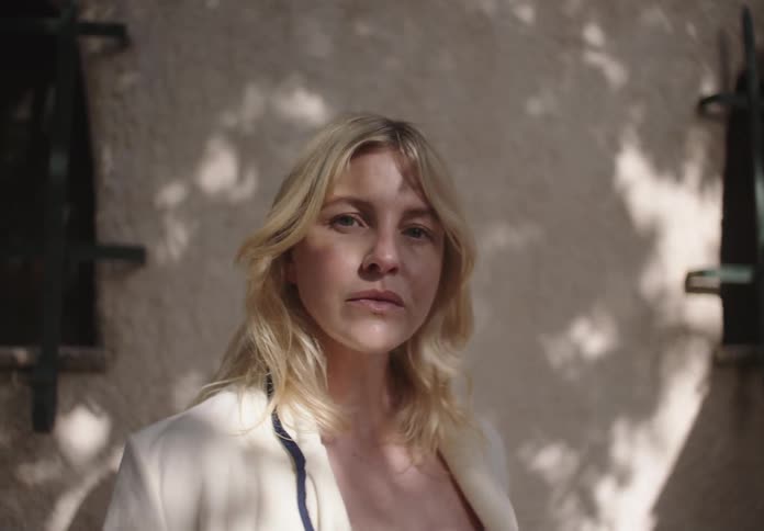 A blonde woman smiles gently while standing in dappled sunlight against a textured wall