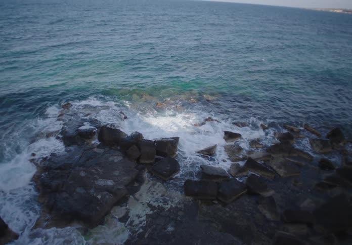 Waves crash against dark coastal rocks beneath a vast open sea