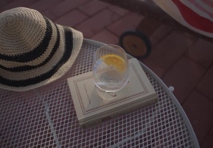 A straw sun hat rests on a patio table next to a book being used as a coaster for a refreshing glass of water with an orange slice