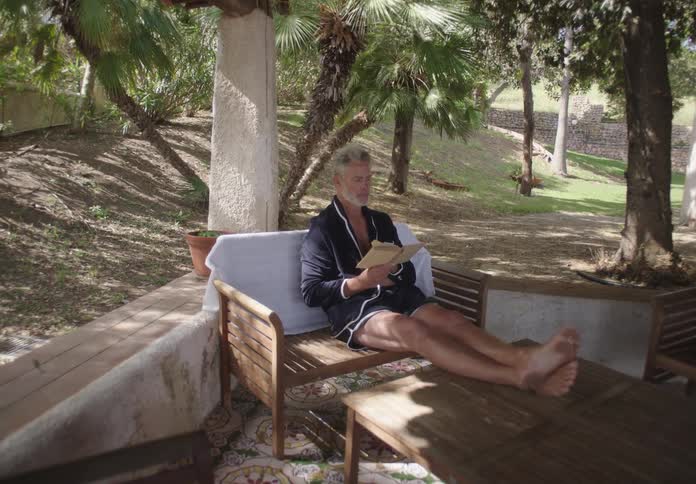 A man relaxes on an outdoor sofa reading a book in a shaded patio surrounded by trees