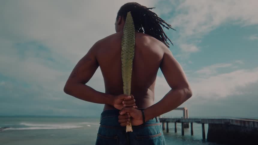 A shirtless man stands at the beach holding a large tropical leaf behind his back facing the ocean