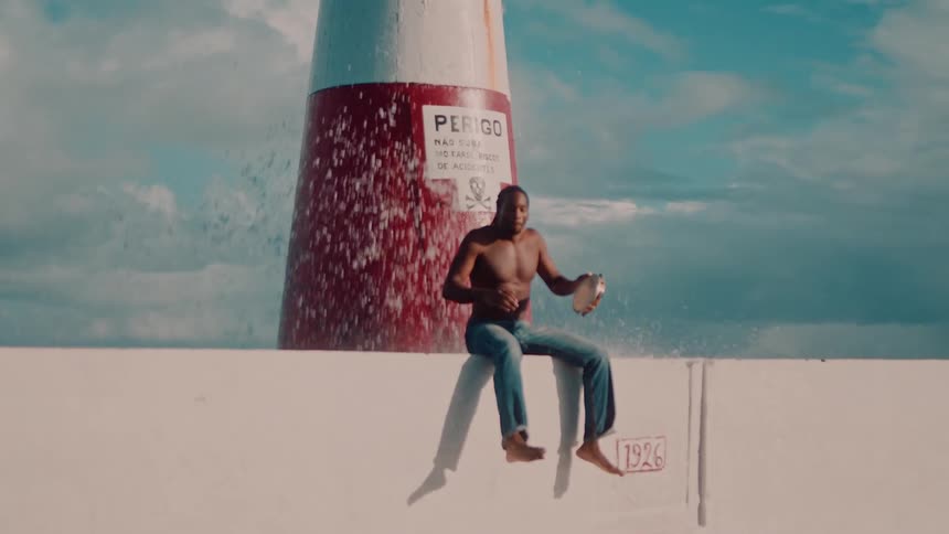 A shirtless man sits barefoot on a white wall smiling as he plays a tambourine with a red and white lighthouse behind him and waves splashing behind under a blue sky