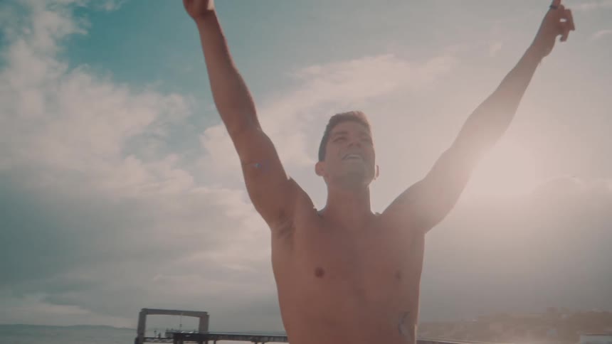 Shirtless man excitedly raises arms while laughing near a pier