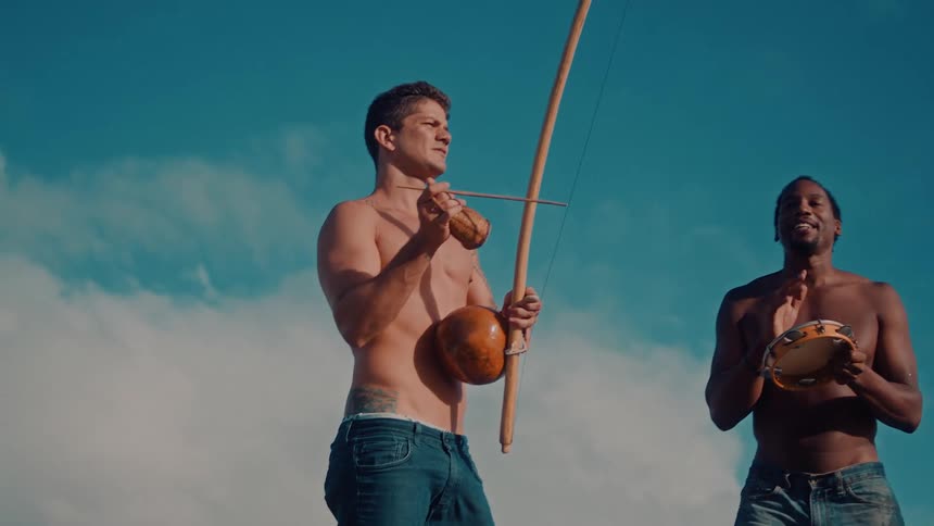 Two shirtless men play traditional brazilian instruments outdoors under a bright blue sky