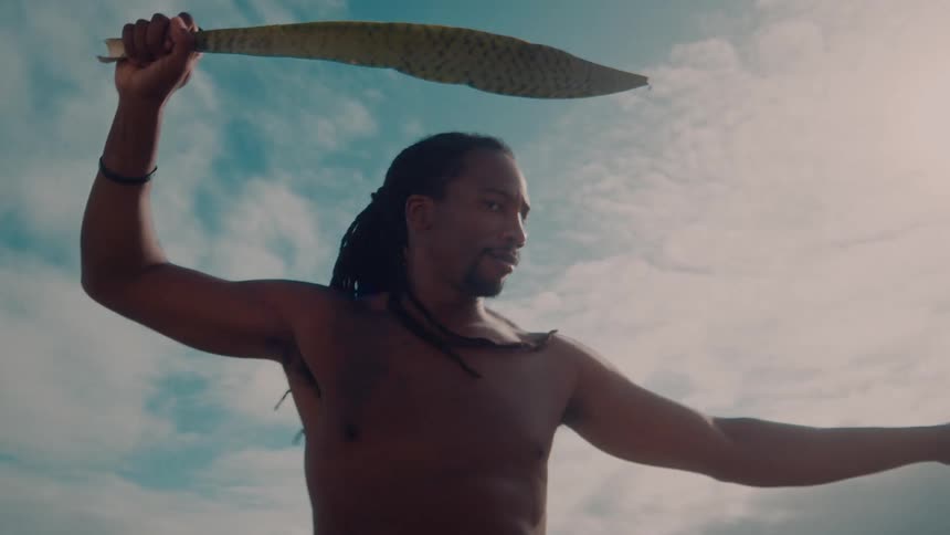 A person stands outdoors beneath a cloudy sky holding a green leaf in one hand and extending the other arm outward