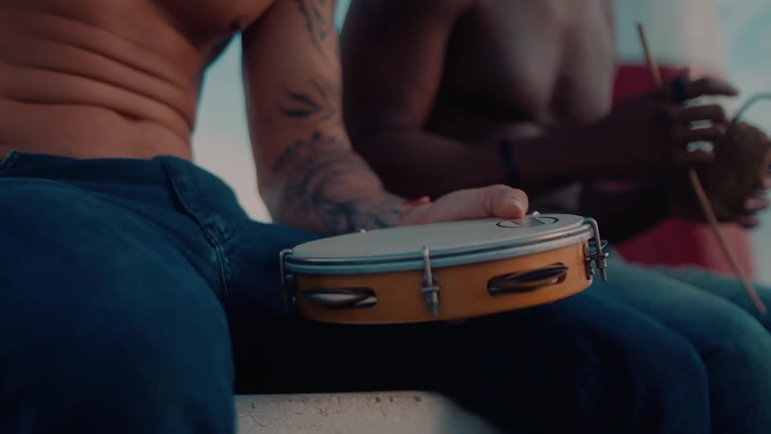 Two shirtless people sit outdoors one holding a tambourine and the other a cowbell