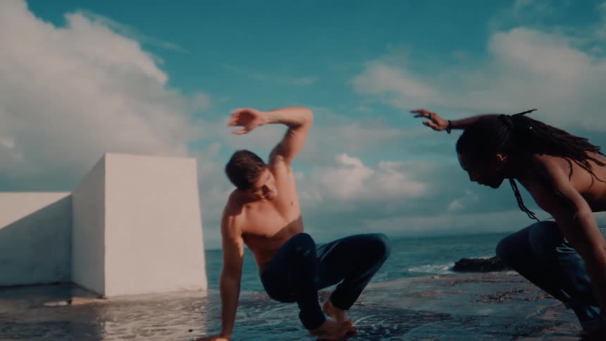 Two men in jeans doing capoeira movements in a seaside setting