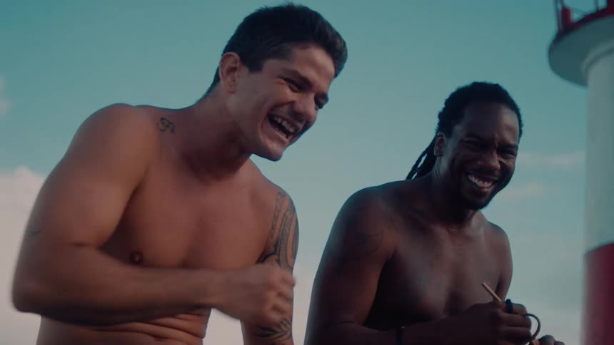 Two shirtless men laugh outdoors as a redandwhite lighthouse peeks into view on the right