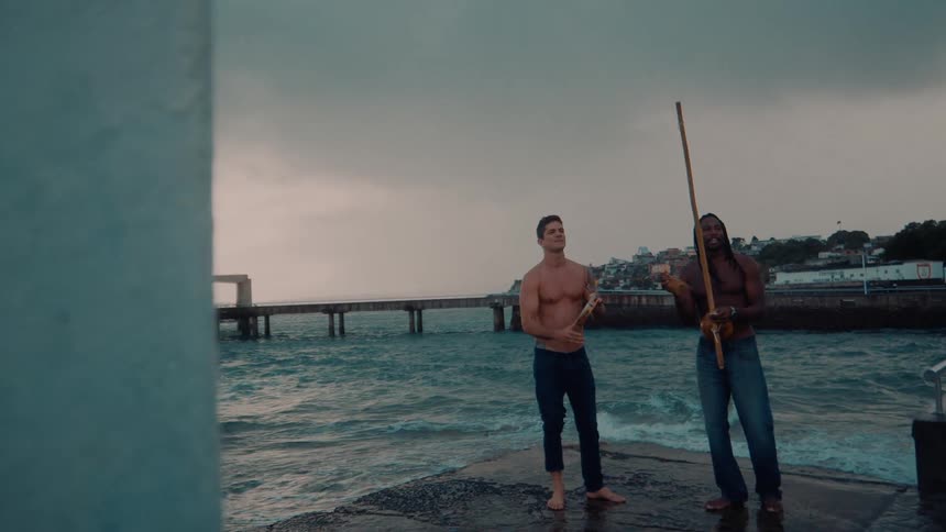 Two men stand at the shore one playing a tambourine and the other holding a long stick as they both sing